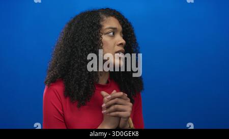 Femme afro-américaine avec les mains serrées près des lèvres dans le studio bleu, les yeux fermés dans l'expression inquiète ; anxiété. Banque D'Images