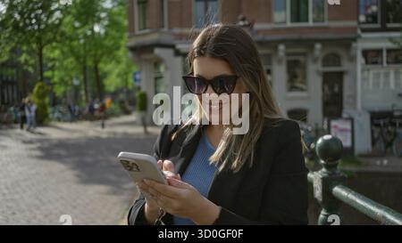 Femme prenant selfie et souriant avec smartphone à amsterdam, marchant le long de canaux pittoresques dans une tenue élégante pendant une journée ensoleillée mettant en vedette l'emblématique c hollandais Banque D'Images