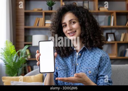 Femme tenant un smartphone avec écran blanc vierge pour maquette, souriant à la caméra et présentant l'application mobile, la nouvelle fonctionnalité ou la promotion tout en étant assis à la maison Banque D'Images