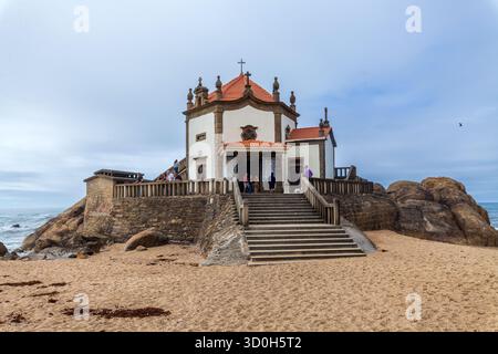 Une chapelle historique avec un toit de tuiles rouges et des murs blancs se trouve au sommet des falaises rocheuses surplombant l'océan, accessible par des escaliers en pierre depuis une plage de sable Banque D'Images