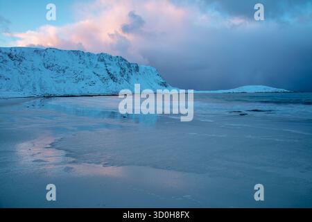 Hiver en Norvège. Soirée sur une plage à Lofoten. Des nuages roses au-dessus des montagnes enneigées se reflètent sur la surface du sable légèrement gelé Banque D'Images