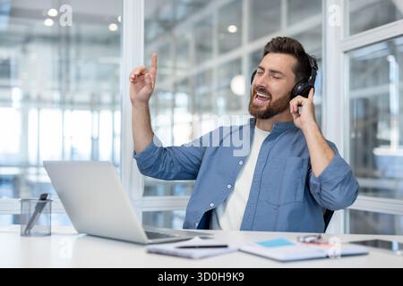 Homme barbu heureux profitant d'une pause de travail, portant des écouteurs et chantant au son de la musique tout en se relaxant à son bureau moderne, trouvant la joie et le soulagement du stress pendant la journée de travail Banque D'Images