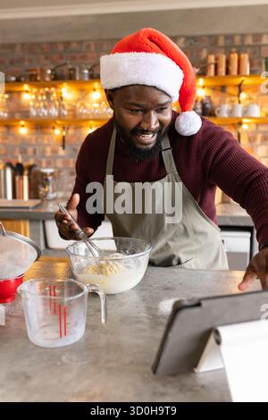 Homme afro-américain adulte moyen portant chapeau de Père Noël fouettant la pâte dans le bol dans la tablette de vérification de cuisine Banque D'Images