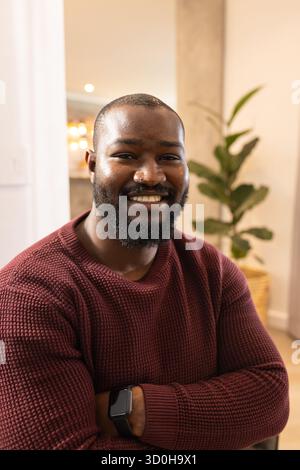 Homme afro-américain assis à la maison portant un pull en tricot Bordeaux et à vérifier smartwatch Banque D'Images