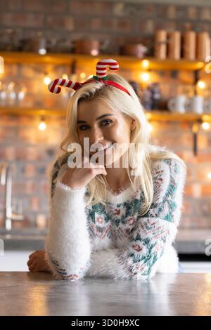 Femme appuyée sur le comptoir lisse dans la cuisine portant un pull de vacances floue et un bandeau de canne de bonbons. Confortable, festif, intérieur, chaleureux, joyeux, célébrez Banque D'Images