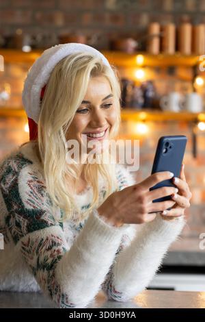 Femme blonde portant chapeau de Père Noël et pull souriant à l'écran de smartphone dans la cuisine par des lumières de chaîne Banque D'Images