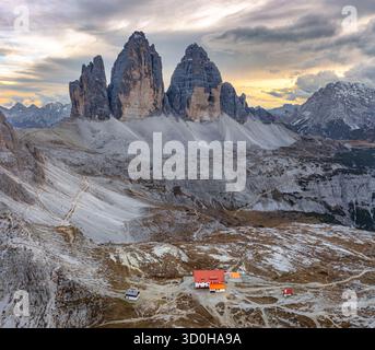Vue aérienne des majestueux sommets Tre Cime di Lavaredo perçant le ciel, avec le Rifugio Locatelli niché en contrebas, baigné dans la douce lueur du crépuscule, en Italie. Banque D'Images