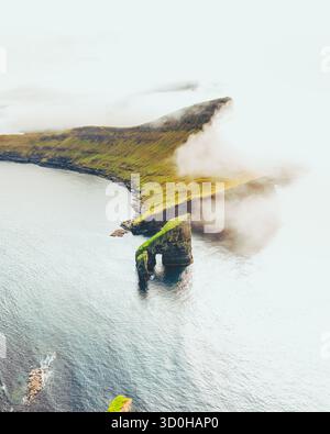 Vue aérienne de la pile marine de Drangarnir perçant à travers des brumes tourbillonnantes, une couronne verdoyante contre le vaste océan scintillant, Drangarnir, îles Féroé. Banque D'Images