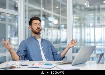 Jeune homme d'affaires méditant à son bureau dans une pose mudra, les yeux fermés, trouvant calme et équilibre travail-vie tout en gérant le stress dans un environnement de travail moderne Banque D'Images
