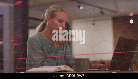 Femme parlant dans une blouse grise tenant smartphone et tapant sur ordinateur portable au bureau avec ordinateur portable Banque D'Images