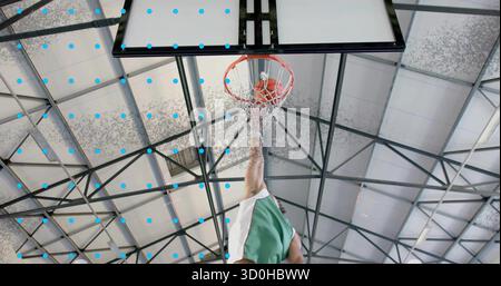 Joueur de basket-ball sautant portant un ballon orange uniforme vert dunking au gymnase, avec fond de filet de jante Banque D'Images