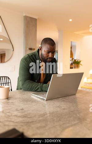 Homme afro-américain penché en avant au comptoir en pierre dans la maison moderne en utilisant un ordinateur portable à côté de la tasse Banque D'Images