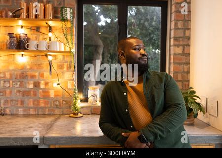 Homme afro-américain appuyé sur le comptoir en béton dans la cuisine rustique avec des lumières de chaîne Banque D'Images