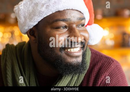 Homme afro-américain souriant portant un chapeau de Père Noël, une écharpe verte et un pull Bordeaux posant à l'intérieur du bar Banque D'Images