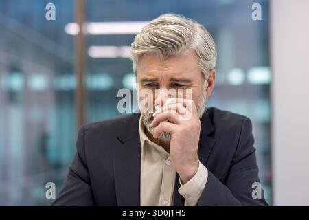 Homme d'affaires mature éprouvant la détresse émotionnelle ou la tristesse, essuyant les larmes avec un mouchoir, transmettant des concepts de deuil, de stress, de perte, d'anxiété et de vulnérabilité dans un cadre d'entreprise Banque D'Images