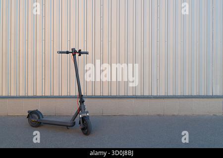 Scooter électrique noir contemporain debout seul sur la chaussée asphaltée à côté d'une clôture en métal ondulé léger sur une journée ensoleillée Banque D'Images