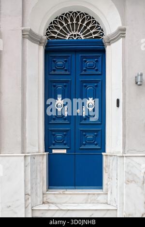 Élégante porte en bois bleu avec panneaux décoratifs, poignées croisées maltaises blanches, fenêtre cintrée avec grille en fer et escaliers en marbre à l'entrée. Banque D'Images