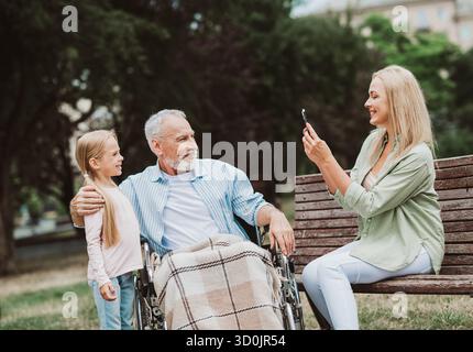 Moment amusant dans un parc familial où la petite-fille filme grand-père dans un fauteuil roulant tandis que maman capture la scène avec chaleur et soin Banque D'Images
