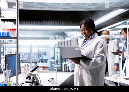 Afro-américaine scientifique travaille dans un laboratoire moderne, menant des expériences avec des équipements de pointe et un ordinateur portable. Axé sur la biologie moléculaire, la biotechnologie et l'innovation dans le domaine de la santé. Banque D'Images