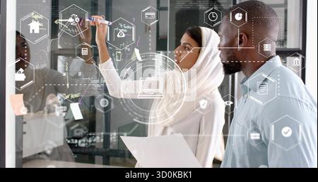 Collaboration de collègues en hijab et chemise annotant un tableau de verre au bureau, avec des icônes holographiques Banque D'Images
