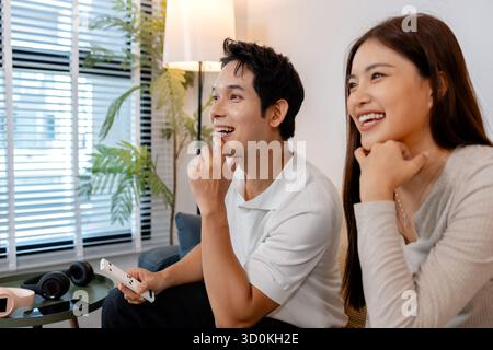 Un jeune couple est assis ensemble sur un canapé, regardant joyeusement la télévision dans un salon confortable. L'homme tient une télécommande tout en souriant et appréciant t Banque D'Images
