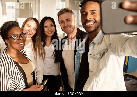 Groupe multiethnique de travailleurs de bureau masculins et féminins souriant joyeusement tout en prenant selfie sur smartphone Banque D'Images
