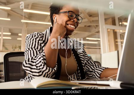 Travailleuse de bureau afro-américaine assise devant un ordinateur portable avec des écouteurs, ayant un chat vidéo et souriant joyeusement, vue à angle bas Banque D'Images