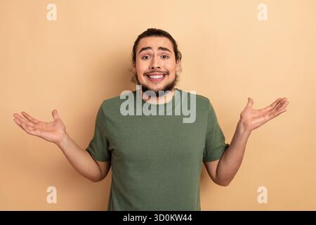 Jeune homme avec un haussement d'épaule et un sourire portant un tshirt vert se tient sur un fond beige dans un moment publicitaire de style de vie décontracté Banque D'Images