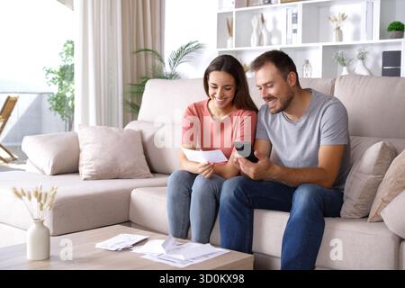 Heureux couple vérifiant le relevé bancaire et téléphone intelligent assis sur un canapé à la maison Banque D'Images