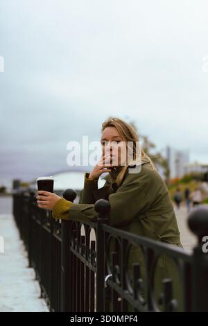 Femme appuyée sur une balustrade noire tenant du café et fumant une cigarette Banque D'Images