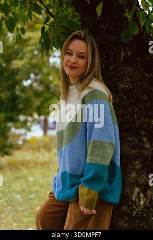 Jeune femme souriante appuyée contre un tronc d'arbre dans un pull coloré Banque D'Images