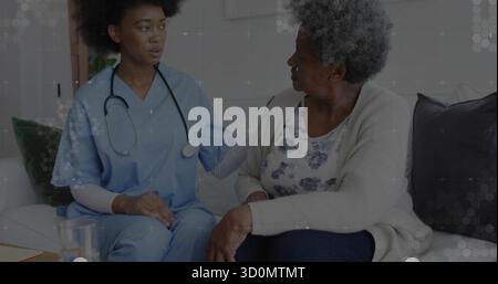 Infirmière portant des gommages bleus réconfortant la femme âgée tout en parlant dans le salon, avec stéthoscope Banque D'Images