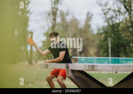 Jeune homme jouant au tennis de table en plein air sur un banc de parc avec une pagaie orange Banque D'Images