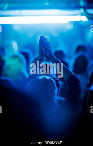 Femme levant les mains et applaudissant parmi le public pendant le concert en direct illuminé avec la lumière bleue de scène. Banque D'Images