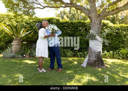 Couple senior embrassant sous l'arbre, tenant le bouquet, célébrant l'amour à l'extérieur. Romance, affection, ensemble, nature, relation, fleurs || autorisation du modèle Banque D'Images