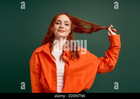 Une jeune femme aux cheveux rouges époustouflants tire ses cheveux de manière ludique tout en vêtue d'une chemise orange. Banque D'Images
