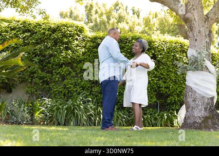 Couple senior dansant joyeusement ensemble dans le jardin ensoleillé, profitant du moment en plein air. Personnes âgées, retraite, bonheur, loisirs, plein air, actif || modèle validé Banque D'Images