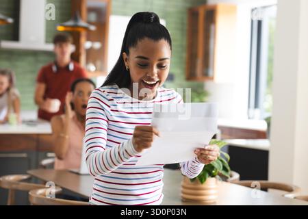 Adolescente célébrant le succès avec des amis dans la cuisine, tenant la lettre excitée. Excitation, célébration, réalisation, amitié, bonheur, émotions || modèle libéré Banque D'Images