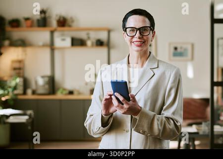 Portrait de femme d'âge moyen souriant tout en utilisant smartphone à l'intérieur Banque D'Images