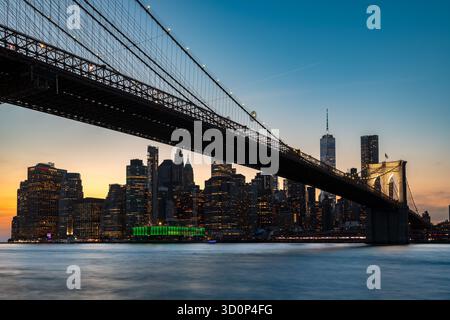 Une vue magnifique sur le pont de Brooklyn enjambant l'eau, avec les gratte-ciel du centre-ville de Manhattan et le quartier financier illuminés au crépuscule. Banque D'Images