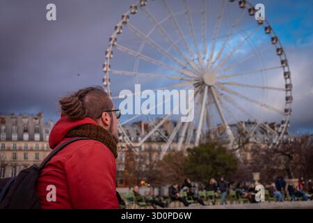 Une personne dans une veste rouge et un sac à dos debout et regardant une grande roue dans un parc urbain animé de Paris, France. Banque D'Images