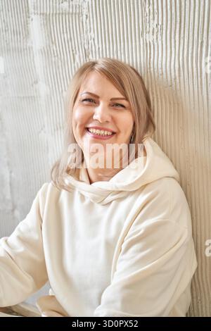 Femme adulte caucasienne souriante aux cheveux blonds portant un pull à capuche appuyé contre le mur blanc texturé. || autorisation du modèle Banque D'Images