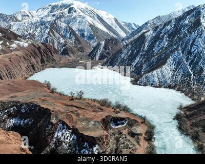 Vue aérienne d'un lac gelé à la base de spectaculaires parois rouges du canyon et massives montagnes enneigées sous un ciel bleu vif Banque D'Images