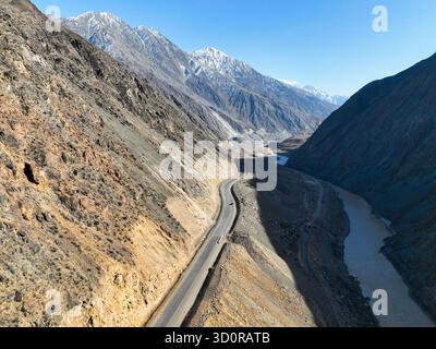 Vue aérienne descendant une gorge de montagne profonde et étroite avec une route sinueuse et une rivière, flanquée de pentes escarpées et accidentées et culminant dans la neige lointaine Banque D'Images