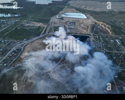 Vue en angle élevé d'un panache de fumée s'élevant d'un site d'enfouissement ou de décharge en feu adjacent à des maisons résidentielles et à un grand bassin de rétention d'eau Banque D'Images
