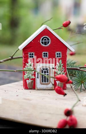 Une charmante maison de jouet rouge, ornée d'arbres de Noël, posée sur une planche de bois, entourée de baies d'hiver et de branches vertes dans une scène fantaisiste Banque D'Images