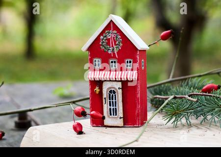 Une charmante maison de jouet en bois rouge ornée d'une couronne festive, évoquant l'esprit de Noël et le charme rustique sur un fond flou. Banque D'Images