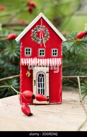 Une charmante scène de Noël : gros plan d'une maison de jouet en bois rouge avec des décorations festives et des hanches roses sur une surface en bois rustique Banque D'Images