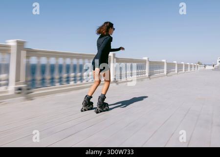 Jeune femme roller le long de la promenade du bord de mer en tenue noire, style de vie actif et concept de liberté Banque D'Images
