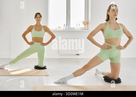 Dans un studio spacieux et bien éclairé, deux femmes effectuent des exercices d'étirement sur tapis. Ils portent des tenues de sport vertes assorties et se concentrent sur leur mouvement Banque D'Images